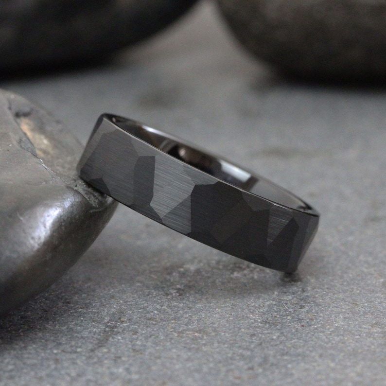 May include: A black wedding band with a geometric patterned design. The band has a matte finish and a slightly rounded edge.