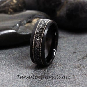 May include: A black tungsten carbide ring with a silver-colored engraved design. The ring has a beveled edge and a polished finish.