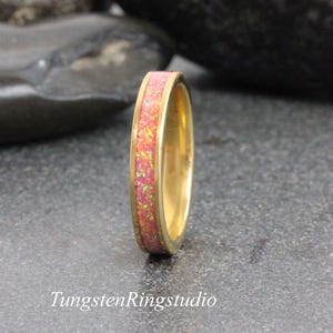 Può includere: Un anello color oro con una fascia di opale rosa e verde. L'anello è posto su uno sfondo scuro. Il testo "TungstenRingstudio" è in basso nell'immagine.