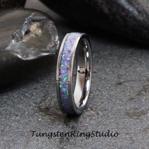 Può includere: Un anello in tungsteno argentato con un'incisione di opale viola e verde. L'anello è lucido e ha un bordo smussato.