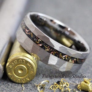 May include: A silver-colored ring with a hammered texture and a dark inlay with copper and gold accents. The ring is next to a bullet casing and a metal tube. The text "TungstenRingStudio" is visible.