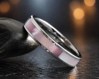 May include: A silver-toned metal ring featuring a smooth, inlaid band of iridescent pink and white shell material. The ring has a polished, reflective finish and a flat profile, showcasing a clean, continuous design around the entire circumference.