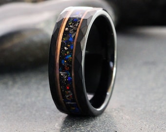 Puede incluir: Un anillo de tungsteno negro con un exterior facetado y una incrustación central de material colorido y brillante. La incrustación está enmarcada por bandas de metal color cobre. El anillo se muestra sobre una superficie gris. El texto "TungstenRingStudio" está en la parte inferior.