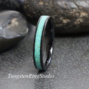 May include: A black ring with a band of shimmering, light green opal. The ring is set against a dark, textured background. The text "TungstenRingStudio" is visible at the bottom.