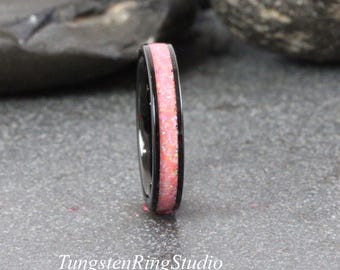 May include: A black tungsten ring featuring a pink opal inlay. The ring has a polished finish and the text "TungstenRingStudio" is visible. The ring is set against a grey surface with dark rocks in the background.