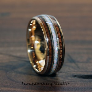 May include: A gold-coloured ring featuring a central band of white opal, framed by dark wood. The ring has a polished finish and a wide band design. The text "TungstenRingStudio" is at the bottom of the image.