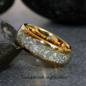 Pu&ograve; includere: Un anello in carburo di tungsteno dorato con un'incisione bianca di meteorite macinata. L'anello &egrave; su una superficie grigia.