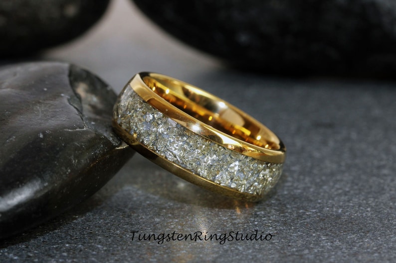 Pu&ograve; includere: Un anello in carburo di tungsteno dorato con un'incisione bianca di meteorite macinata. L'anello &egrave; su una superficie grigia.