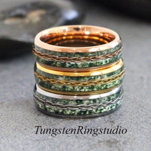 May include: A stack of four rings with a green and white speckled inlay and braided metal bands. The top ring is rose gold, the second is gold, and the bottom ring is silver. The text "TungstenRingstudio" is at the bottom.