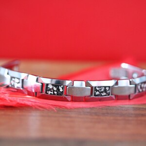 May include: A silver bracelet with black accents. The bracelet is made of metal links and has a modern design.