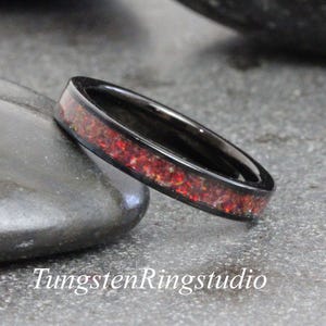 Può includere: Un anello in tungsteno nero con una fascia di materiale intarsiato rosso e arancione. L'anello è posto su uno sfondo grigio. Il testo "TungstenRingstudio" è visibile in basso nell'immagine.