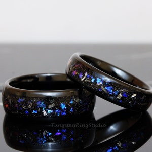 May include: Two black tungsten carbide rings with a galaxy-inspired design. The rings are embedded with blue, silver, and black glitter.