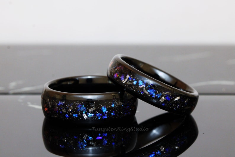 May include: Two black tungsten carbide rings with a galaxy-inspired design. The rings are embedded with blue, silver, and black glitter.
