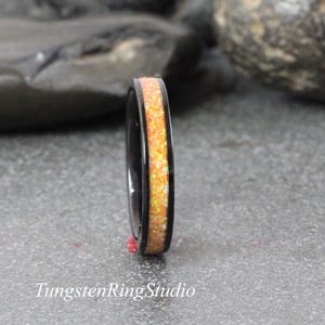 Può includere: Un anello nero con una fascia centrale di materiale arancione e giallo simile all'opale. L'anello è posto su una superficie grigia con il testo "TungstenRingStudio" in basso.
