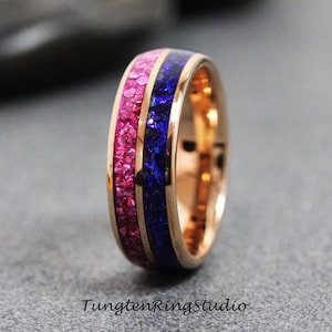 May include: Rose gold ring with a pink and blue inlay. The ring features a band of pink stones and a band of blue stones, set in a polished rose gold band. The text "TungstenRingstudio" is at the bottom of the image.