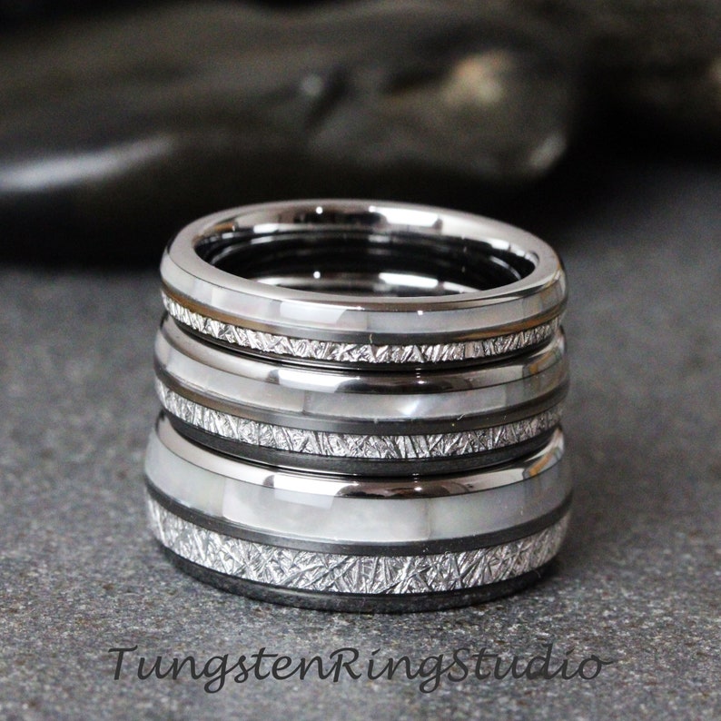 May include: Stack of four silver tungsten carbide rings with a white inlay and a brushed finish. Each ring has a different pattern of lines etched into the surface.