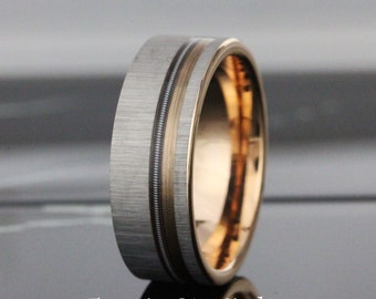 Puede incluir: Un anillo de tungsteno plateado con una incrustación de oro rosa y un acabado cepillado. El anillo tiene un diseño acanalado alrededor de la incrustación.