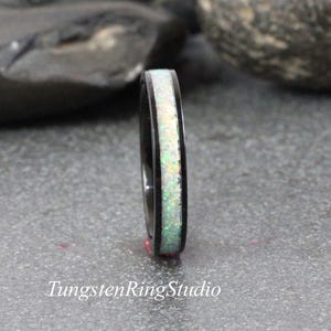 Può includere: Un anello nero con un intarsio di opale bianco. L'opale presenta macchie verdi e gialle. L'anello è posto su uno sfondo grigio. Il testo "TungstenRingStudio" è in basso nell'immagine.