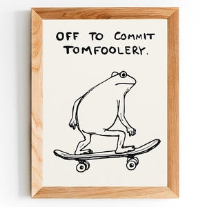 May include: A framed print with a light wood frame. The print features a black and white illustration of a frog on a skateboard, with the text "OFF TO COMMIT TOMFOOLERY" above the frog. The background is off-white.