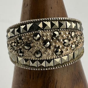 Op de afbeelding: Een zilverkleurige ring met een brede band. De ring heeft drie rijen decoratieve elementen: twee rijen piramidevormige studs en een centrale rij kleine, ronde, getextureerde accenten. De ring wordt getoond op een houten standaard.