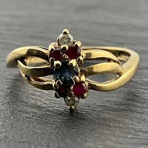May include: A gold ring featuring a central dark blue sapphire flanked by four small red rubies and two clear white diamonds arranged in a vertical cluster. The band consists of two split gold strands that meet at the sides of the setting.