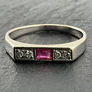 May include: A silver-coloured metal ring featuring a central rectangular-cut pink gemstone flanked by two small round clear stones on each side, set within a smooth, polished band that tapers slightly towards the back of the ring.