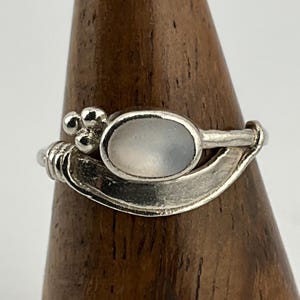 May include: A silver ring with a pale oval stone. The ring has a decorative design with a cluster of small beads and a curved, leaf-like element. The ring is displayed on a wooden stand.