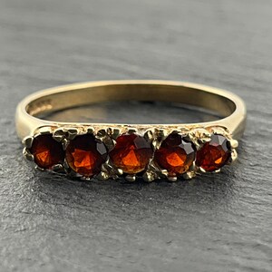 May include: A gold ring featuring five round, faceted, deep red garnet gemstones set in a row. The band is smooth and polished, while the setting around the stones has a textured, organic appearance. The ring is crafted from yellow gold.