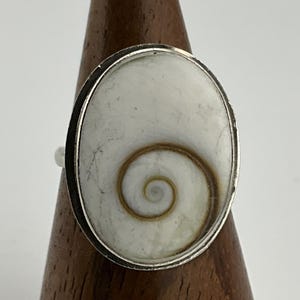 May include: Silver ring with an oval white shell and a brown spiral pattern. The shell is set in a silver bezel. The ring is displayed on a wooden ring holder. The shell has a smooth, polished surface.