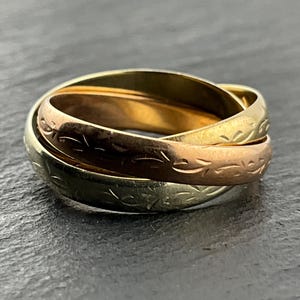 Puede incluir: Un anillo de tres bandas entrelazadas fabricado en metal con tonos amarillo, rosa y blanco. Cada banda individual presenta un delicado patrón grabado de hojas o enredaderas que se repite a lo largo de toda la circunferencia del anillo.