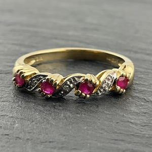 May include: A gold ring featuring a twisted band design with four round-cut red rubies set at intervals. Between each ruby, the band incorporates small, clear accent stones set into a silver-toned metal channel, creating a textured appearance.