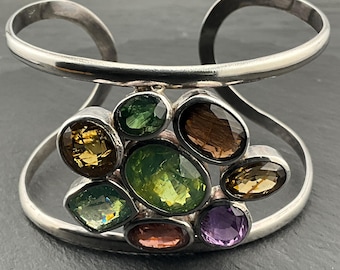 May include: A silver cuff bracelet with a cluster of colourful gemstones. The bracelet has a wide, open design with a curved top and a central section adorned with various oval and teardrop-shaped stones in shades of green, brown, yellow, and purple.
