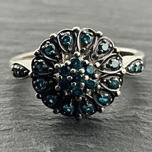 Puede incluir: Un anillo de tono plateado con un diseño circular de racimo. El centro está compuesto por pequeñas piedras redondas de color azul oscuro, rodeadas por un halo de engastes en forma de lágrima, cada uno con una piedra azul oscuro. La banda también tiene piedras azules.