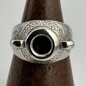 May include: A silver ring with a circular black centre, featuring an ornate floral design around the band. The ring has two small, curved extensions on either side. The ring is displayed on a wooden stand.