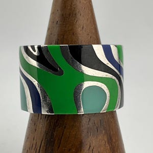 Peut inclure: Une large bague colorée avec un design abstrait audacieux. La bague présente un motif de formes vertes, noires, bleues et turquoise soulignées d'argent. La bague est présentée sur un support en bois.