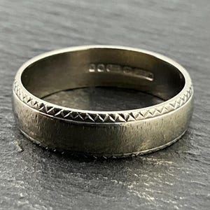 Puede incluir: Un anillo plateado con una banda texturizada y un borde decorativo. El anillo tiene un diseño simple y clásico, adecuado para el uso diario. La banda del anillo tiene aproximadamente 0,64 cm de ancho.