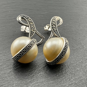 Pode incluir: Um par de brincos de prata com uma gota de pérola e um design de marcasita. Os brincos têm uma forma delicada e curva.