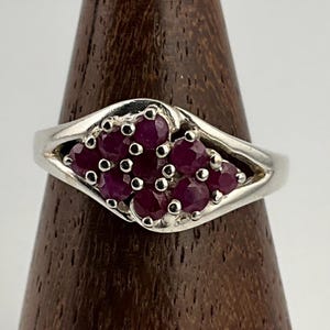 May include: A silver ring featuring a cluster of deep red gemstones. The gemstones are set in a decorative pattern, creating a focal point. The ring is displayed on a wooden stand, showcasing its design and craftsmanship.