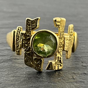Könnte beinhalten: Ein Goldring mit einem runden, facettierten grünen Peridot-Edelstein in der Mitte. Die Ringschiene und die Fassung weisen eine texturierte, unregelmäßige Oberfläche mit vertikalen, gezackten Metallelementen auf, die den Stein umgeben.