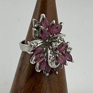 May include: A silver ring with a floral design. The ring is set with oval-cut, deep pink gemstones and small, clear accent stones. The ring is displayed on a wooden stand against a white backdrop.