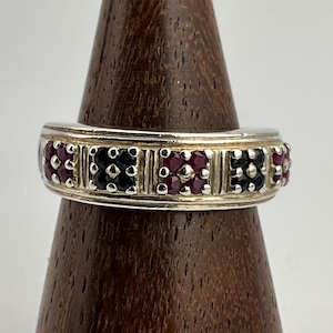 Könnte beinhalten: Ein silberner Ring mit abwechselnden Abschnitten aus roten und schwarzen Edelsteinen. Der Ring wird auf einem hölzernen Ringhalter präsentiert, der sein detailliertes Design und den Farbkontrast hervorhebt. Die Edelsteine sind in einem sich wiederholenden Muster angeordnet.