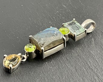 Puede incluir: Un colgante de plata con una piedra de labradorita gris grande, una piedra de peridoto verde más pequeña y una piedra de aguamarina azul claro. El colgante tiene una anilla de plata y una cadena de plata.