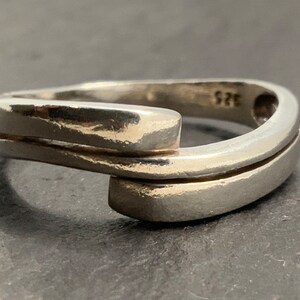 Pode incluir: Um anel de prata com um design moderno e único, apresentando duas bandas paralelas que se cruzam em ângulo. O anel é marcado com o número "925", indicando que é feito de prata de lei.