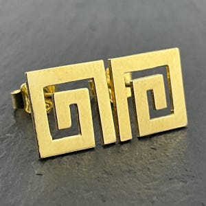 May include: A pair of gold-toned stud earrings featuring a square-shaped Greek key pattern. Each earring has a flat, polished surface with a cutout design that forms a continuous geometric meander motif. The earrings include standard post backs.