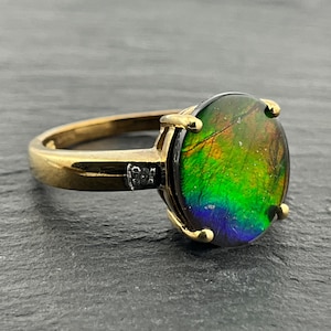 Puede incluir: Un anillo de oro con una piedra preciosa de ammolita redonda e iridiscente en tonos vibrantes de verde, amarillo y morado. La piedra está sujeta por un engaste de cuatro garras, con una pequeña piedra de acento redonda y clara a cada lado.