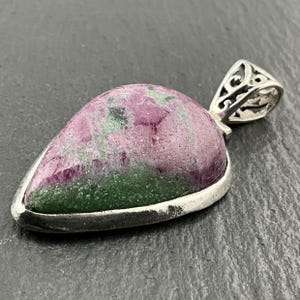 May include: A teardrop-shaped pendant with a polished ruby zoisite stone. The stone features a mix of pink, green, and white colours, set in a silver-coloured metal frame. The bail has an ornate, openwork design.