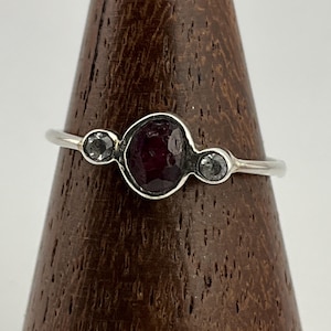 Peut inclure: Une bague en argent ornée d'une pierre précieuse rouge foncé, ovale et facettée, flanquée de deux petites pierres grises rondes. La bague est présentée sur un support en bois, mettant en valeur son design élégant et son savoir-faire.