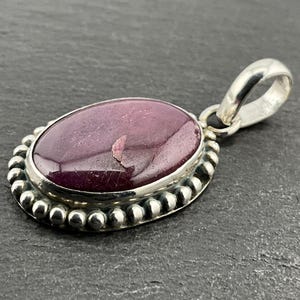 May include: Oval pendant featuring a deep purple gemstone set in a silver bezel. The pendant has a beaded border and a silver loop for a chain. The gemstone has a slightly uneven surface.