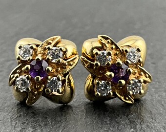 Könnte beinhalten: Ein Paar goldfarbene Ohrstecker. Jeder Ohrring hat ein Blumenmuster mit klaren und violetten Edelsteinen. Die Ohrringe sind auf einer dunkelgrauen Oberfläche platziert.