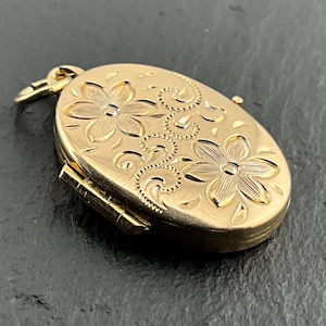 May include: An oval gold locket featuring an engraved floral pattern with two prominent six-petalled flowers and swirling scrollwork details. The piece includes a small hinged bail at the top and a side hinge mechanism for opening the locket.